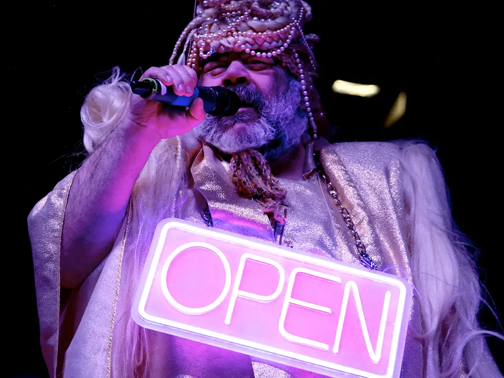 Ein Mann in einem rosanen Kostüm mit Mikrofon und eine Neon-Schild mit der Aufschrift OPEN